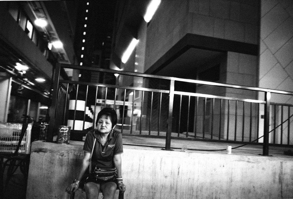Waterloo Street, Singapore. Shooting at the late hours, you don’t get many people around. This lady is obviously drawn out after a long day, serving customers at the adjoining food center. Here in Singapore, we have food centers (also called “coffee shops” or “hawker centers”) where small food vendors set up stalls in a large area with communal sitting. A good portion of these places are open 24-hours a day, seven days a week, to cater to the city’s populace. I took this with a 21mm lens, thus I had to get up close for this shot. She didn’t have the patience for me, as you can tell from the skewed lines but I think it adds to the photo.